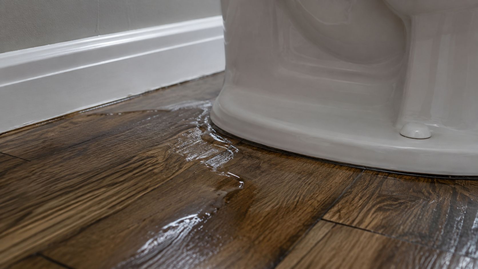 A close-up shot showing water leaking from the base of a white toilet onto a hardwood floor.