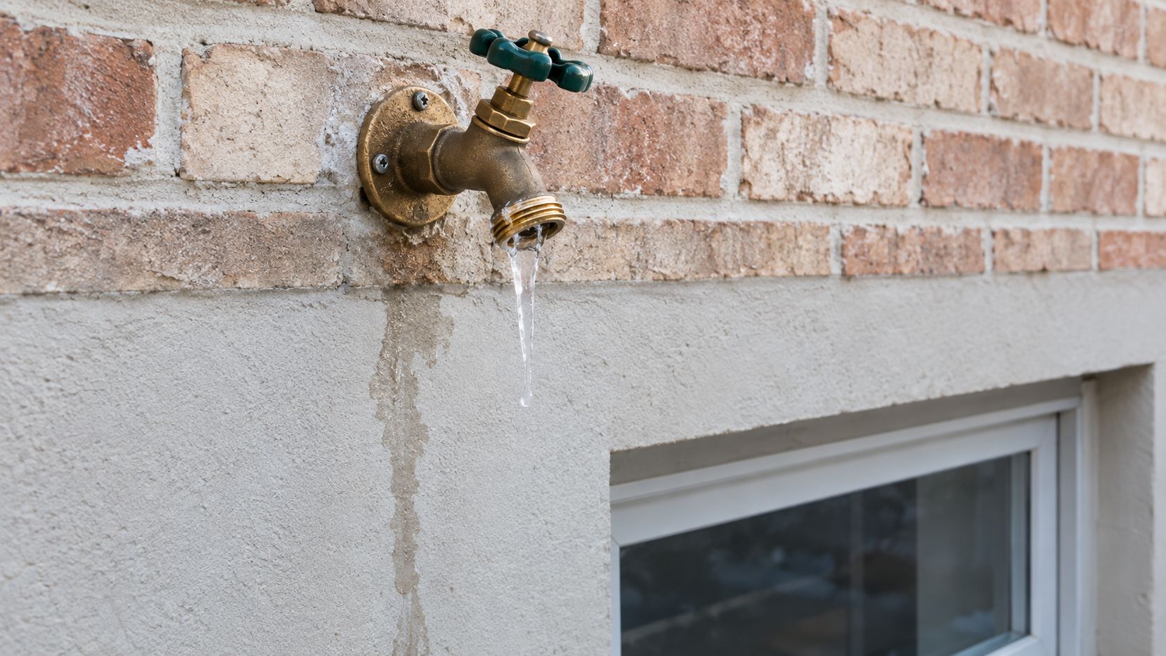 An outdoor water faucet leaking and freezing with icicles forming on the nozzle against a brick wall.